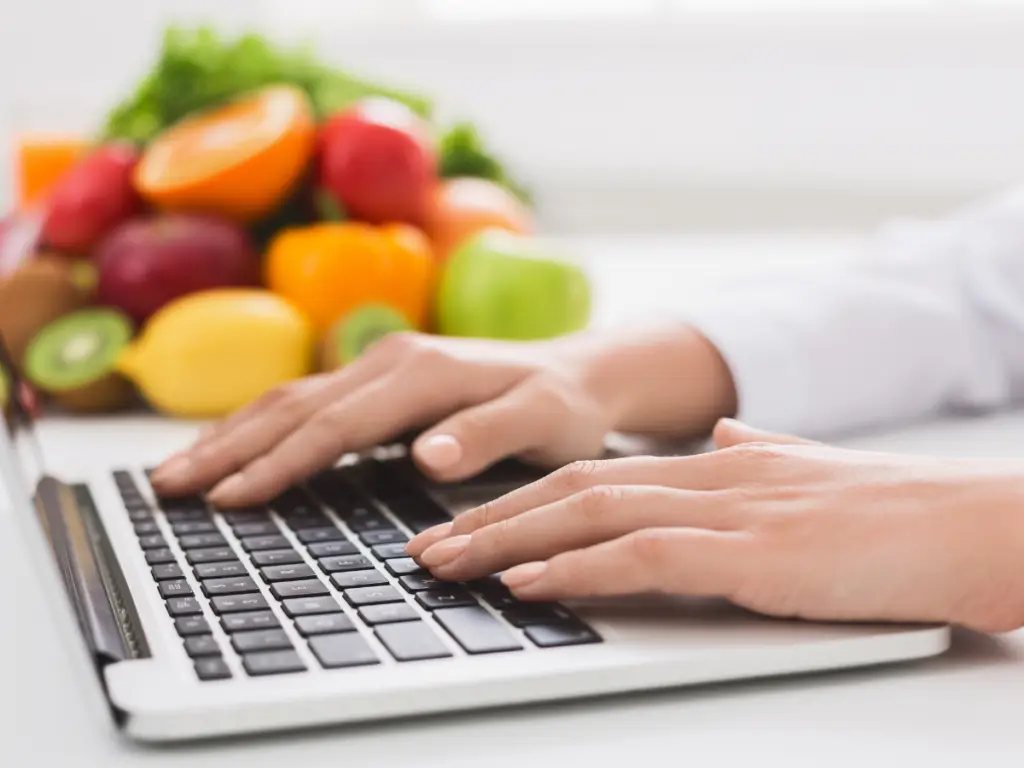 Typing on a laptop with fresh fruits and vegetables in the background - Nutrition research and planning